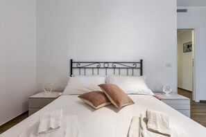 A neatly made bed with white linens and two brown pillows, a bedside table with a white lamp, and a white wall with a door leading to another room.