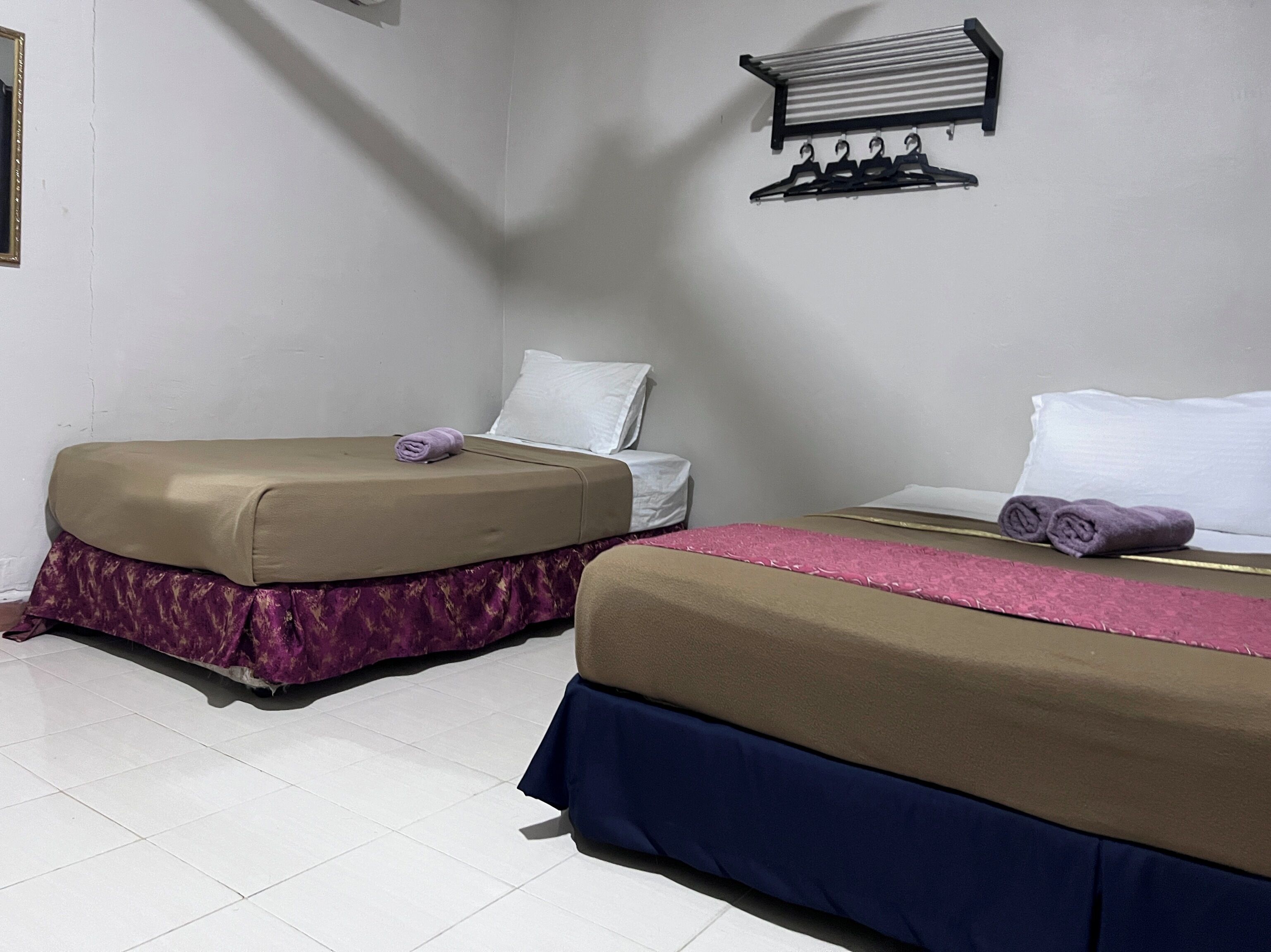 A hotel room with two beds, each with a different colored bedspread and a rolled towel on one. There is a wall-mounted shelf with hangers above the beds.