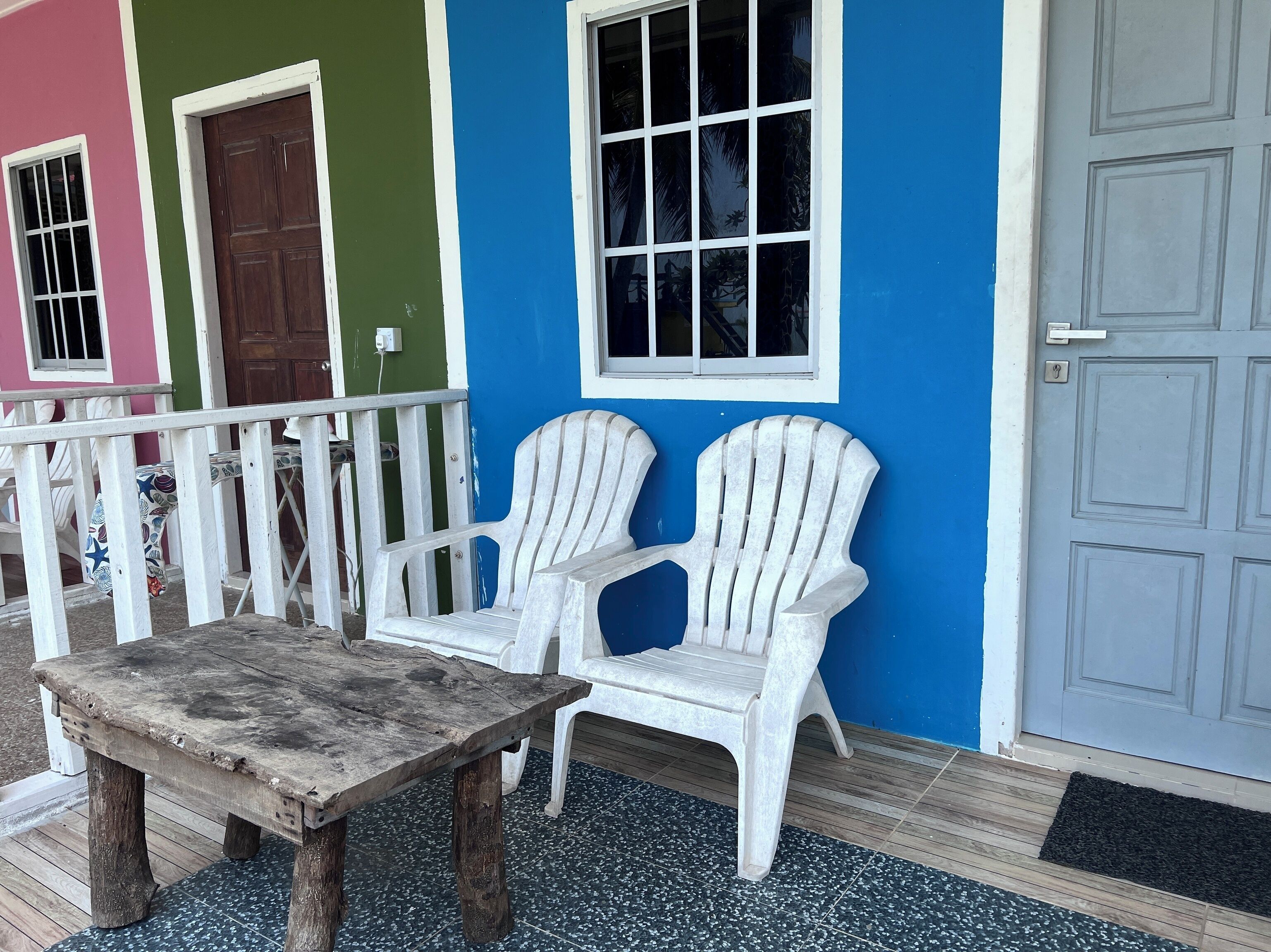 A porch with a wooden table and two white Adirondack chairs, a green door, and a blue wall with a window.