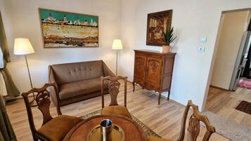 A living room with a sofa, wooden cabinet, and a dining table with chairs. There are two lamps, a painting on the wall, and a potted plant on the cabinet.