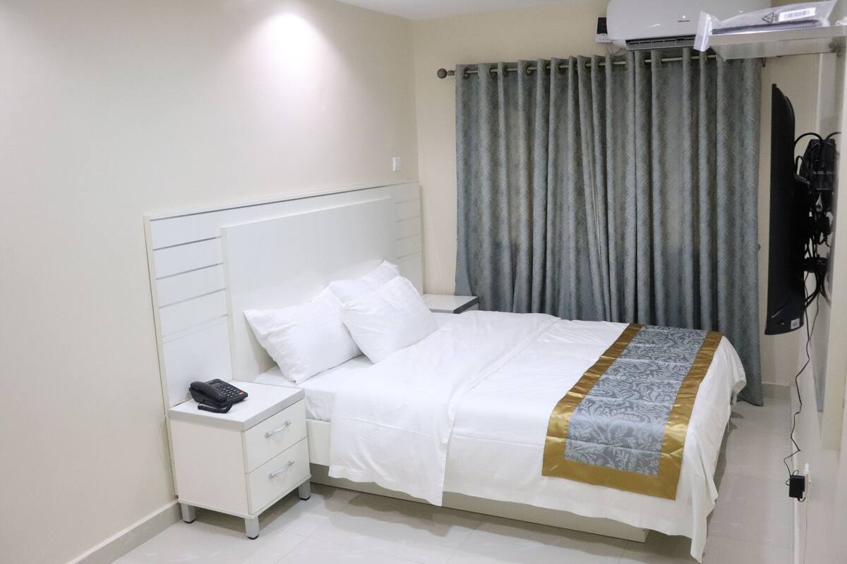 A hotel room with a white bed, a bedside telephone, a flat-screen TV mounted on the wall, and a window with grey curtains.