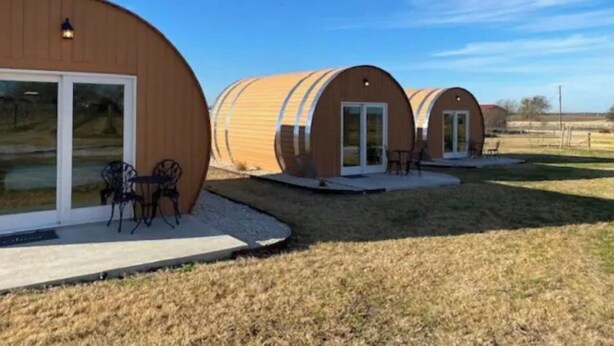 A row of cylindrical, wooden cabins with sliding glass doors, each featuring a small patio area with outdoor seating. - Riesling  Cabin (Seguin)