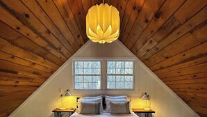 Un dormitorio con techo inclinado, una cama grande, dos lámparas de noche y un ventanal con vistas a árboles.