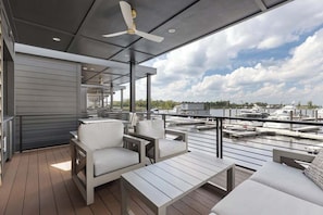 Terraza al aire libre techada con mobiliario, ventilador de techo y vista a un puerto deportivo.