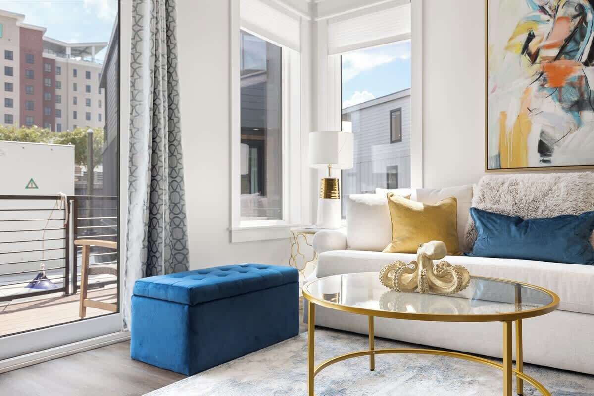 A modern living room with a white sofa, blue ottoman, and a glass coffee table.