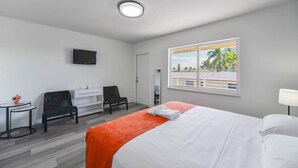 Habitación de hotel con cama, escritorio, silla, televisión y ventana con vista a palmeras.