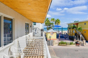 A balcony with a wooden ceiling overlooking a beach area with palm trees and a blue umbrella.