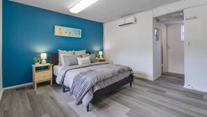 A bedroom with a bed, bedside tables, a nightstand, a wall-mounted air conditioner, and a door leading to another room.