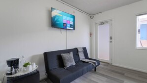 A modern living room with a flat-screen TV mounted on the wall, a dark sofa with cushions, a small side table with a coffee machine and cups, and a door with a window to the right.