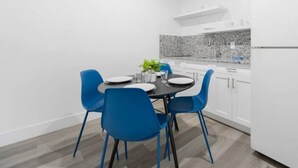 A modern kitchen with white cabinets, a granite backsplash, and a black countertop. There is a dining table with blue chairs and a small potted plant.