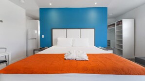 Una habitación de hotel con una cama grande, ropa de cama blanca, una colcha naranja, una pared de acento azul, un armario blanco y un escritorio con una silla.
