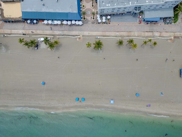An aerial view of a beach with palm trees, umbrellas, and a building with a blue roof.