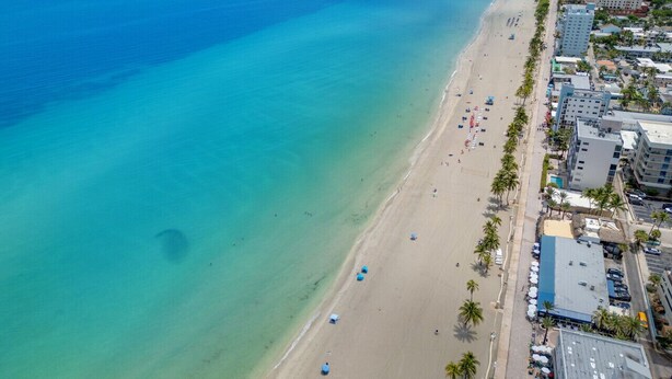 Eine Vogelperspektive zeigt einen Strand mit klarem türkisblauem Wasser, einem Sandufer und angrenzende Gebäude mit Balkonen, die auf den Strand blicken.