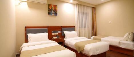 A hotel room with three single beds, a wooden headboard, a bedside table with a phone, and an air conditioning unit on the wall.