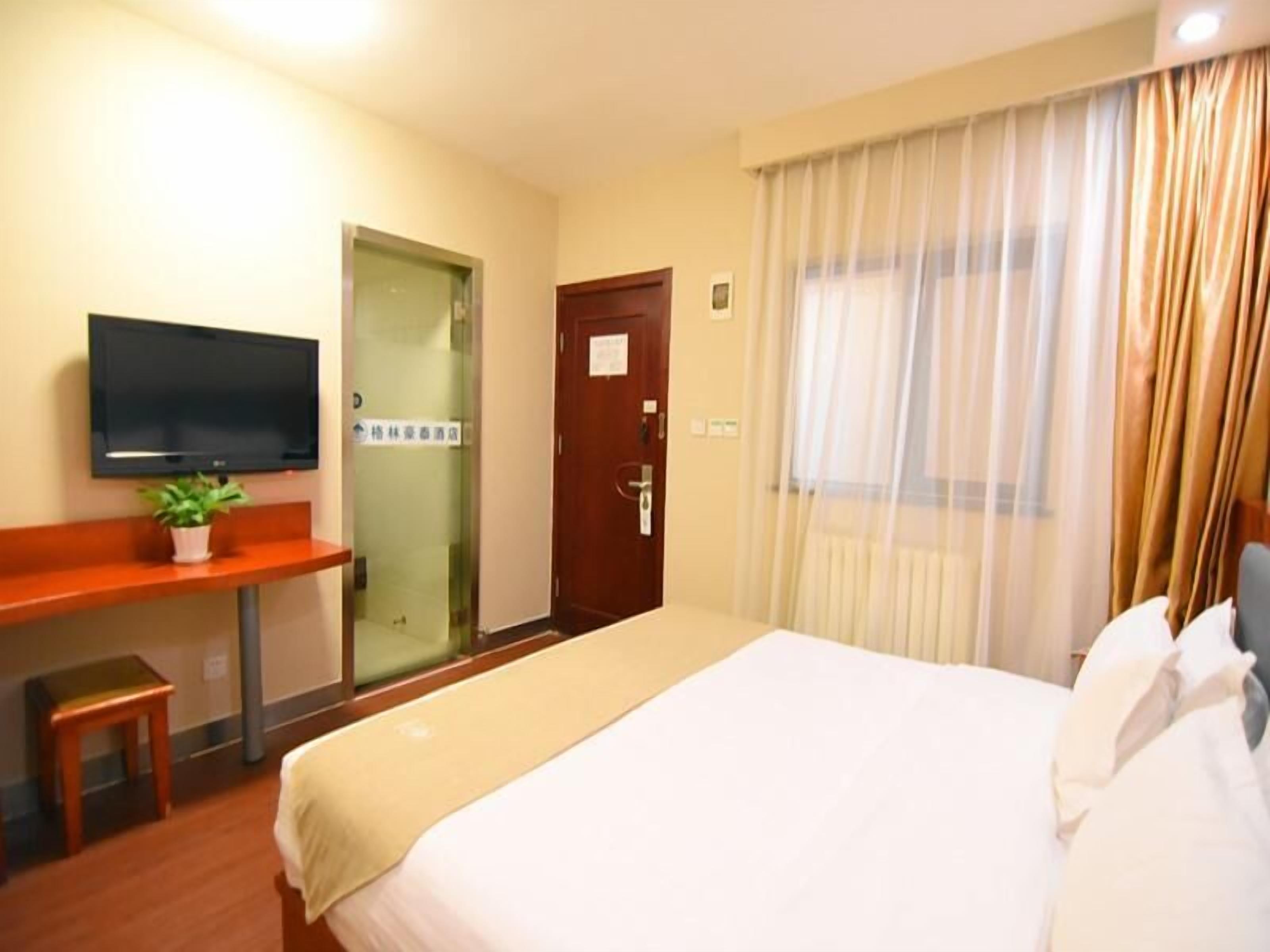 Habitación de hotel con cama, televisor en un soporte, escritorio con planta, puerta y ventana con cortinas.