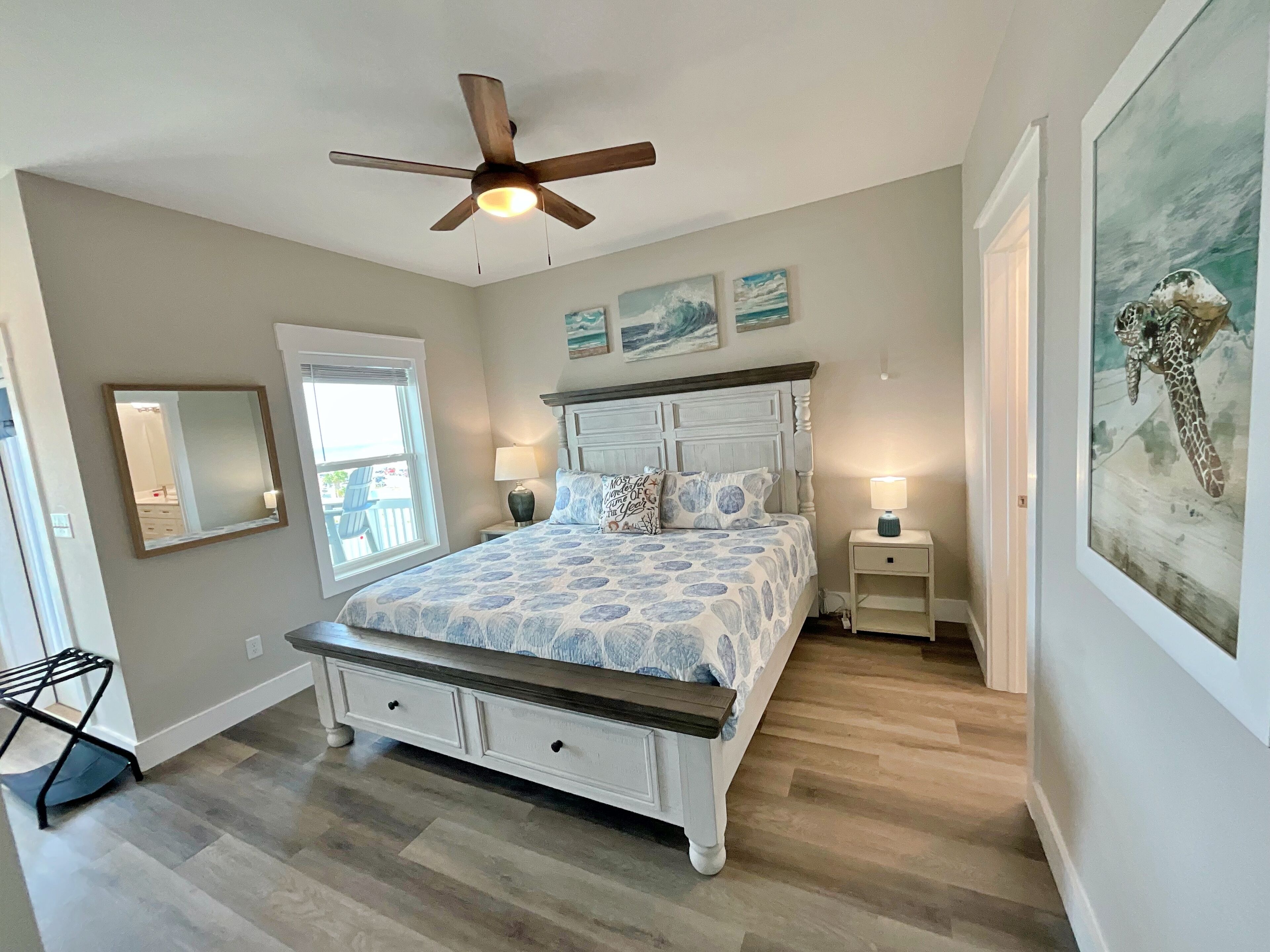 A bedroom with a bed, a ceiling fan, a window, a mirror, a nightstand, and a painting.