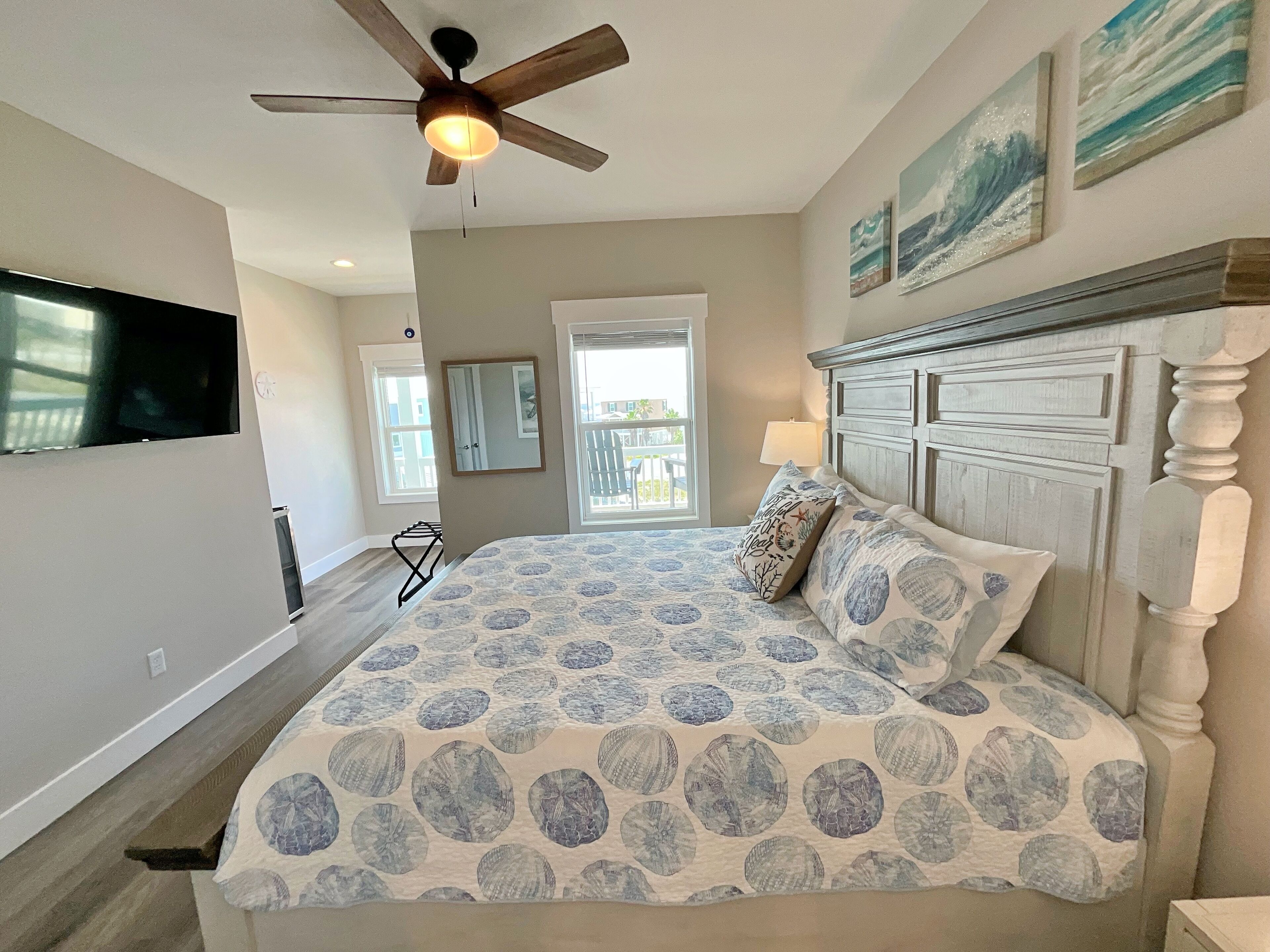 A bedroom with a bed, a ceiling fan, a TV mounted on the wall, and a window with a view of the outside.