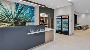 Un área de recepción moderna con un mural digital, un mostrador de recepción, una zona de estar y un refrigerador con bebidas.