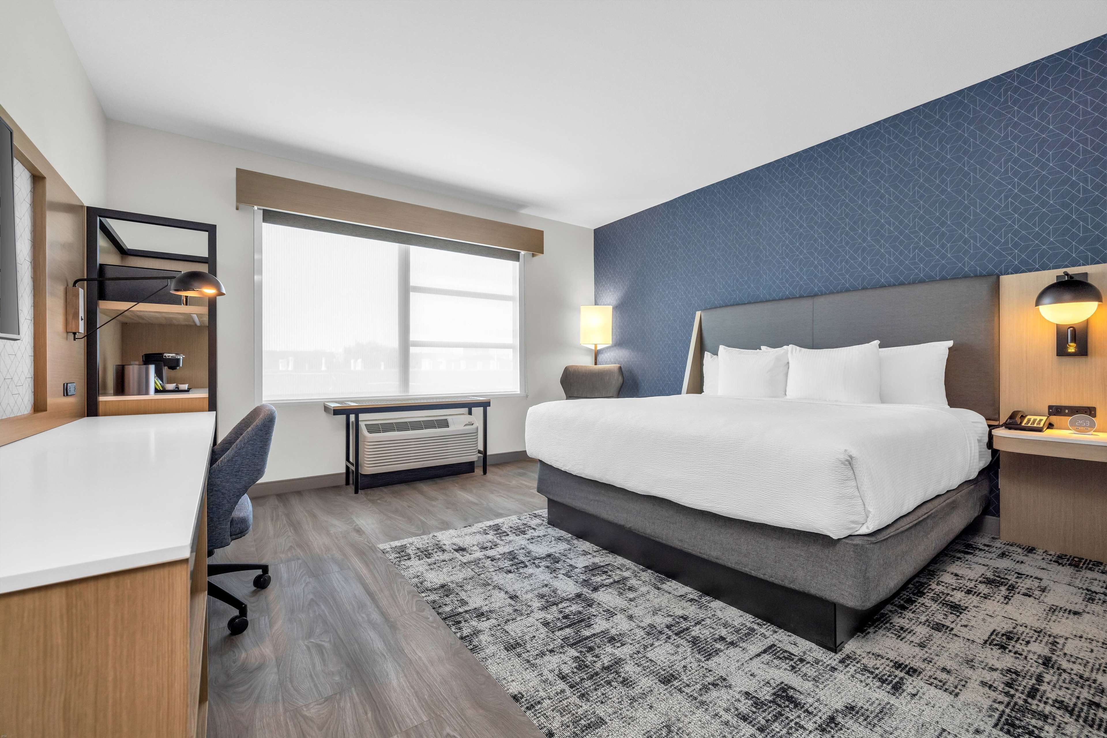 A hotel room with a large bed, a desk with a chair, a desk lamp, a nightstand with a lamp, and a window with blinds.