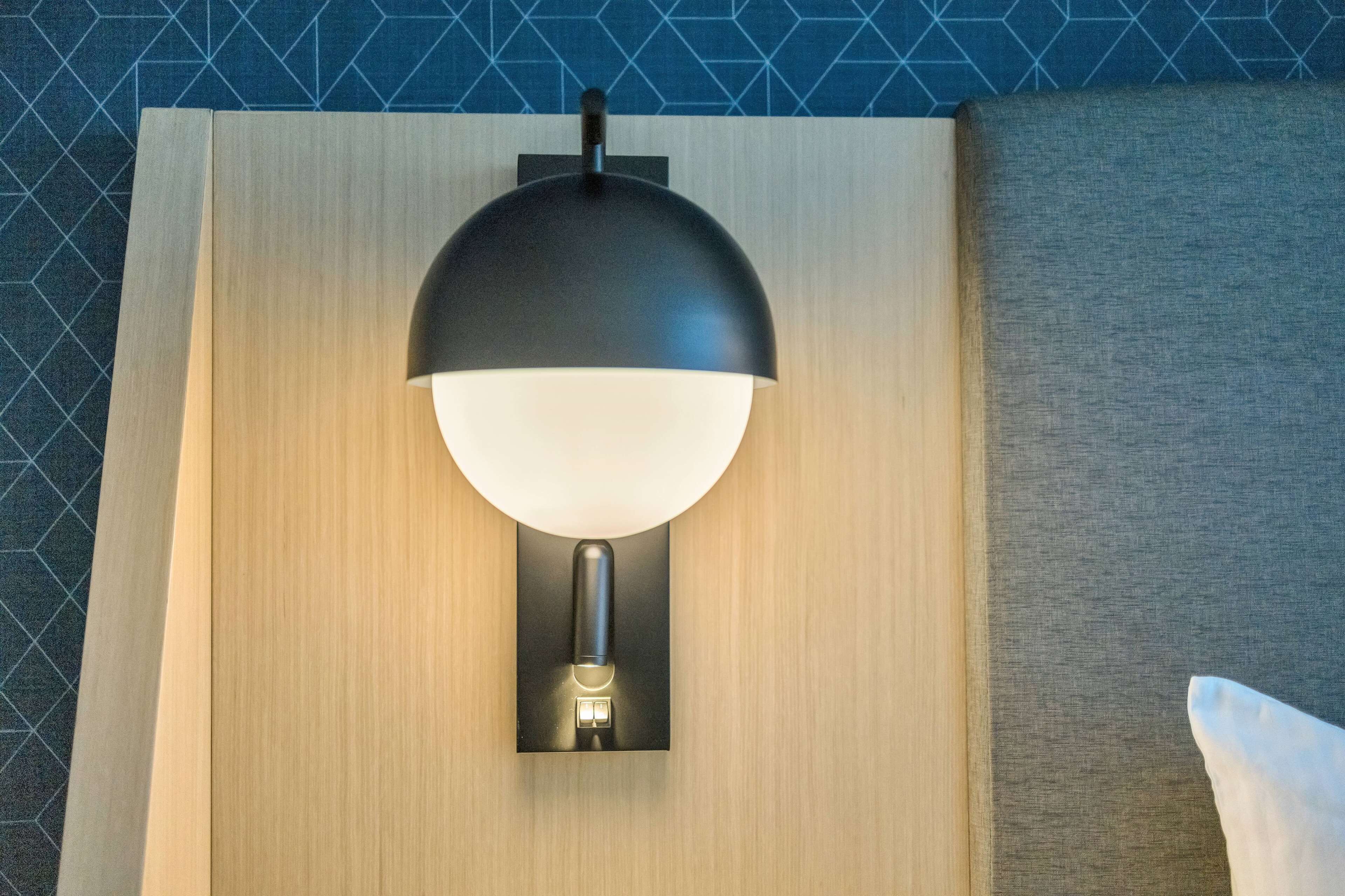 Uma luminária moderna fixada na parede, com cúpula preta e base branca, montada em um painel de madeira com papel de parede geométrico ao fundo.