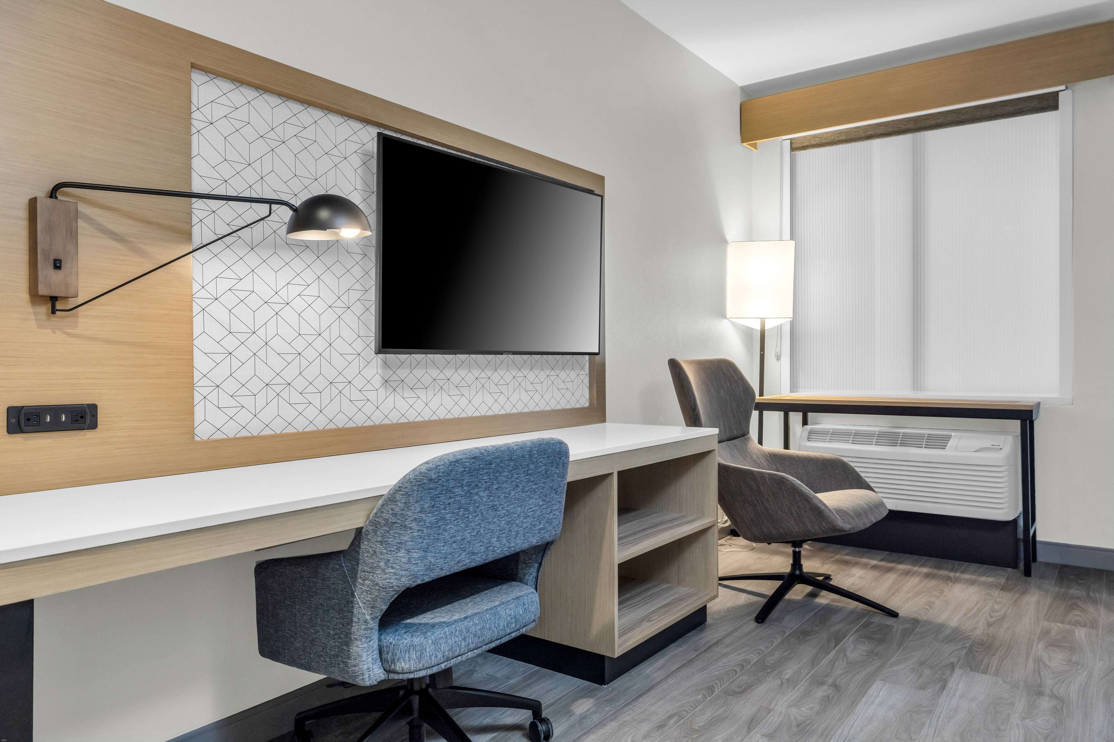 Quarto de hotel moderno com uma televisão de tela plana grande presa a uma parede com padrão geométrico, uma escrivaninha com duas cadeiras, um abajur e uma janela com persianas.