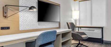 Quarto de hotel moderno com uma televisão de tela plana grande presa a uma parede com padrão geométrico, uma escrivaninha com duas cadeiras, um abajur e uma janela com persianas.