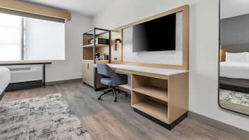 A modern hotel room with a large bed, a desk with a TV, a chair, and built-in shelving.