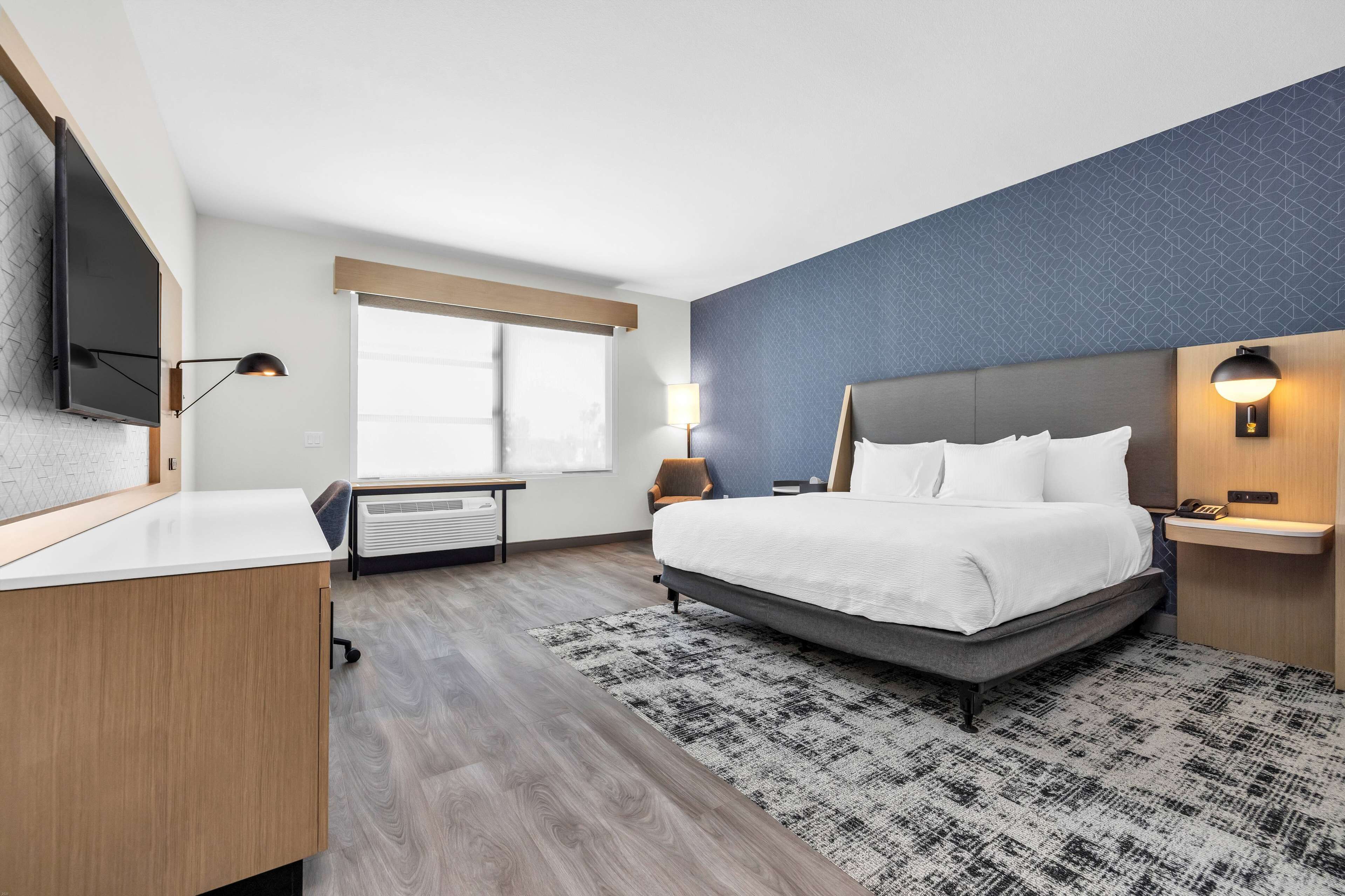 A modern hotel room with a large bed, a desk, a flat-screen TV, and a window with blinds.