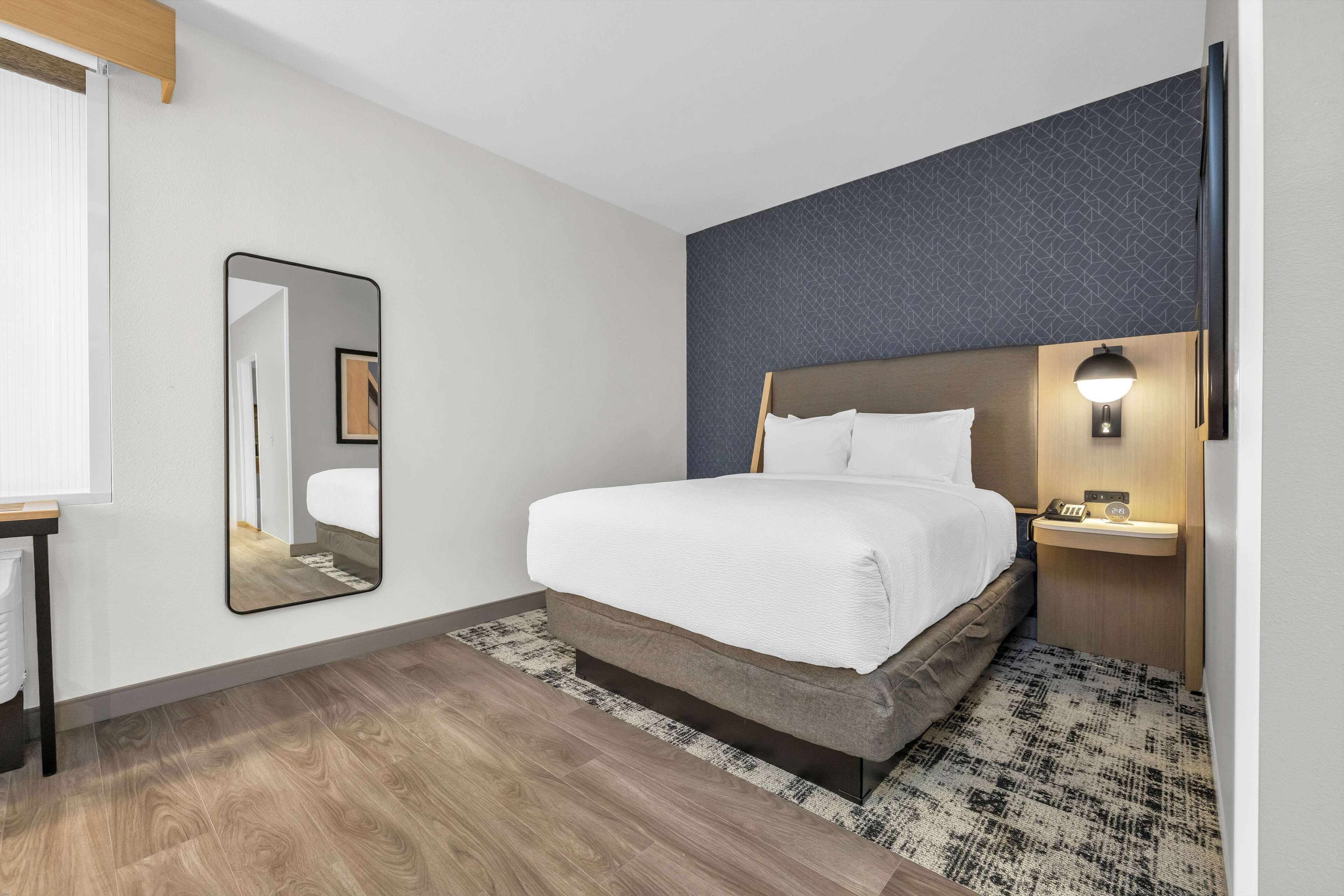 A modern hotel room with a large bed, a bedside table, a nightstand, a mirror, and a wall-mounted TV.