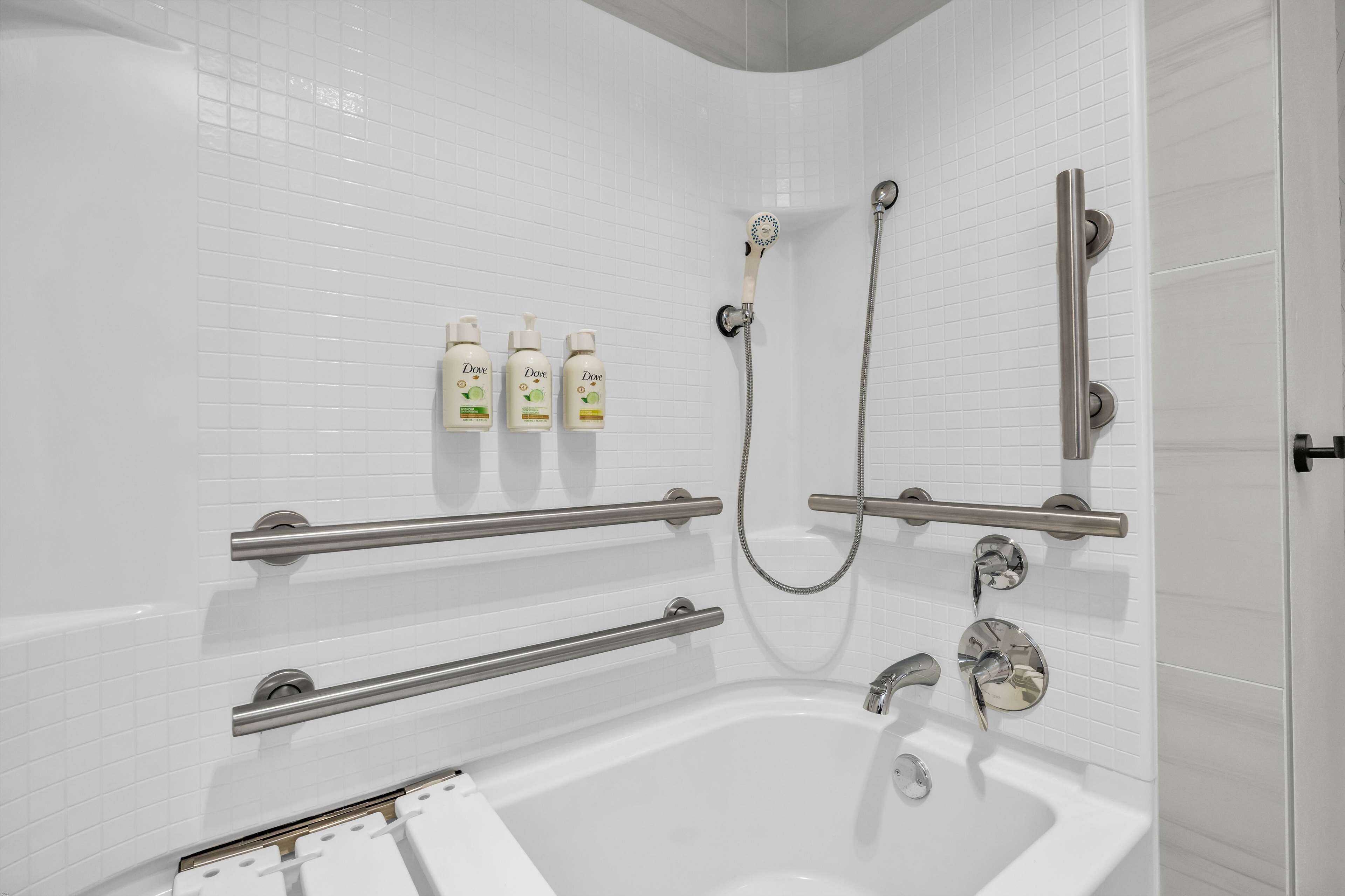 A modern bathroom with a white tiled wall, a bathtub, a shower with a handheld showerhead, and three bottles of Dove body wash.