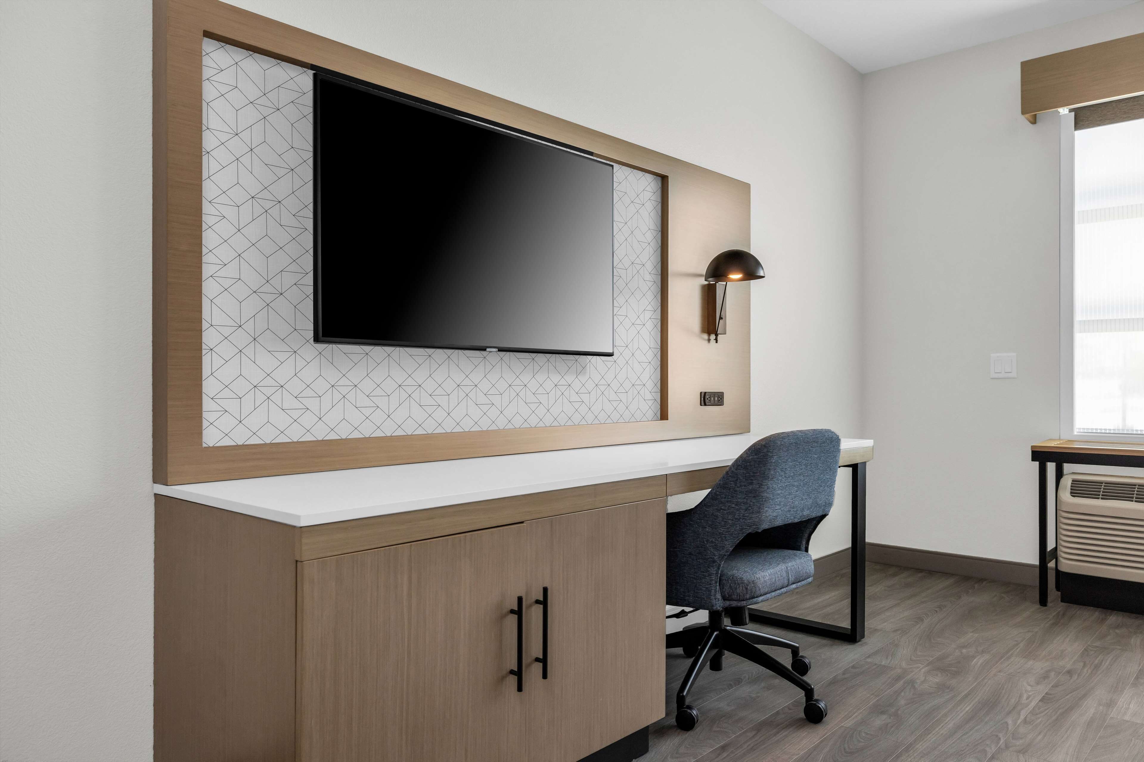Un espace de bureau moderne doté d’un grand téléviseur à écran plat fixé à un mur orné d’un motif géométrique, d’un bureau avec une chaise et d’une lampe fixée au mur.