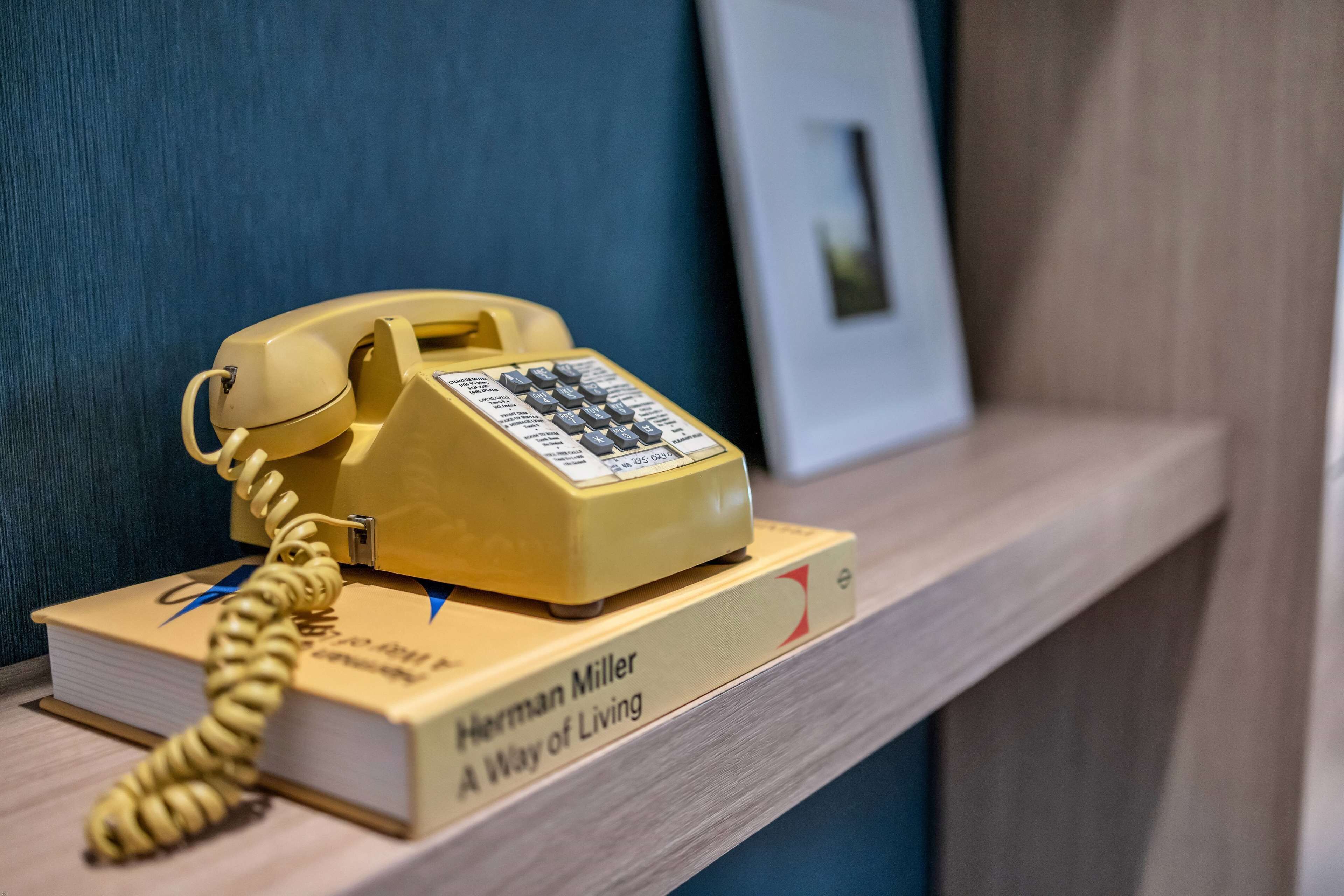 A yellow rotary phone on a wooden shelf with a book titled "A Way of Living" by Herman Miller.