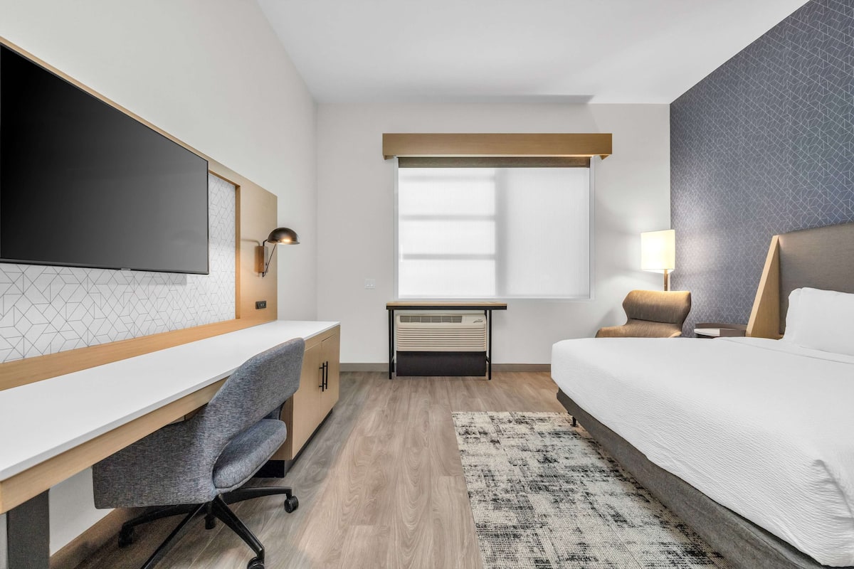Une chambre d’hôtel moderne dotée d’un grand lit, d’un bureau avec une chaise, d’un téléviseur à écran plat, d’une fenêtre avec des stores et d’un tapis à motifs.