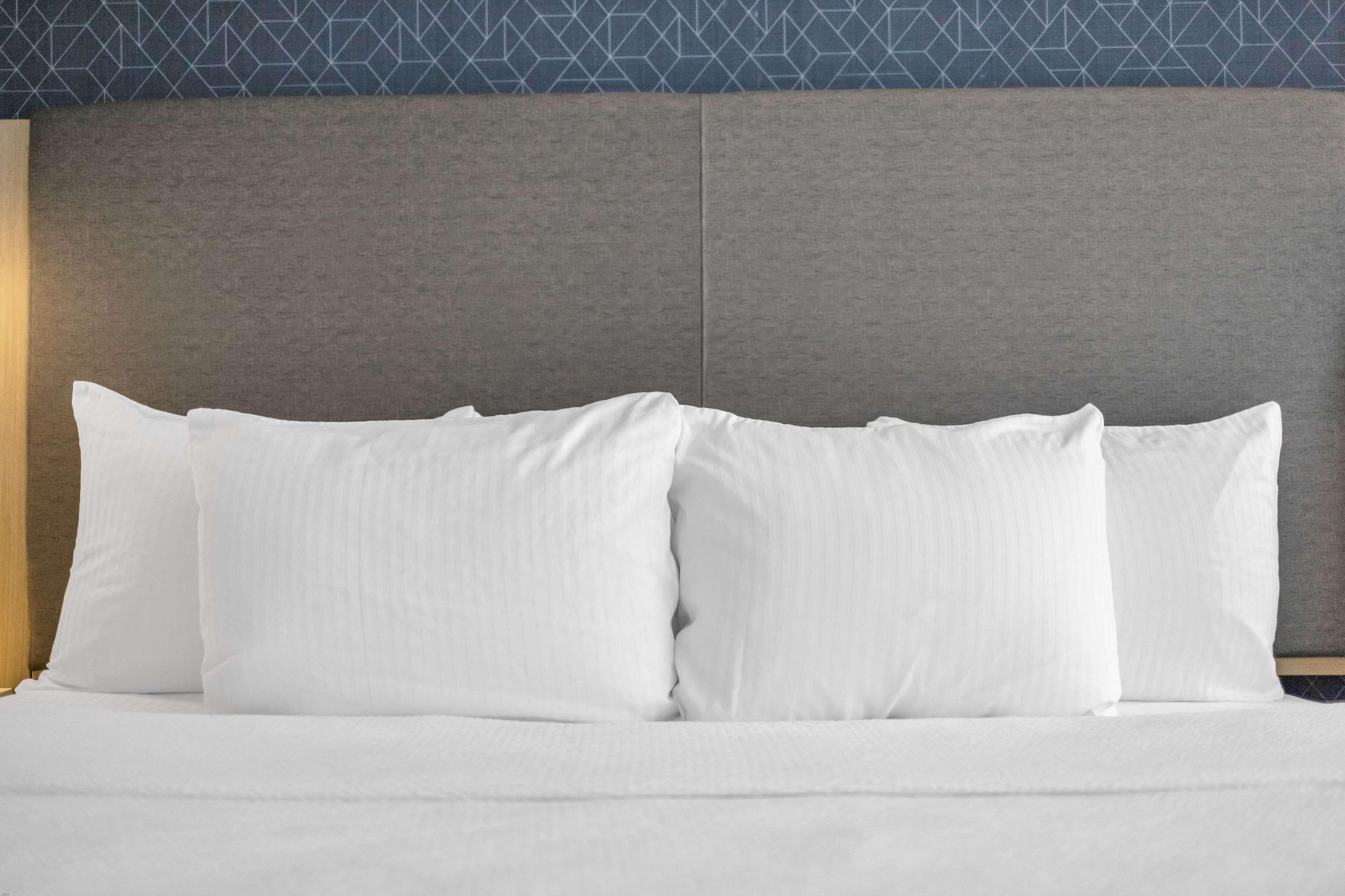 Una cama con ropa de cama y almohadas blancas, cabecera gris y un papel tapiz estampado al fondo.
