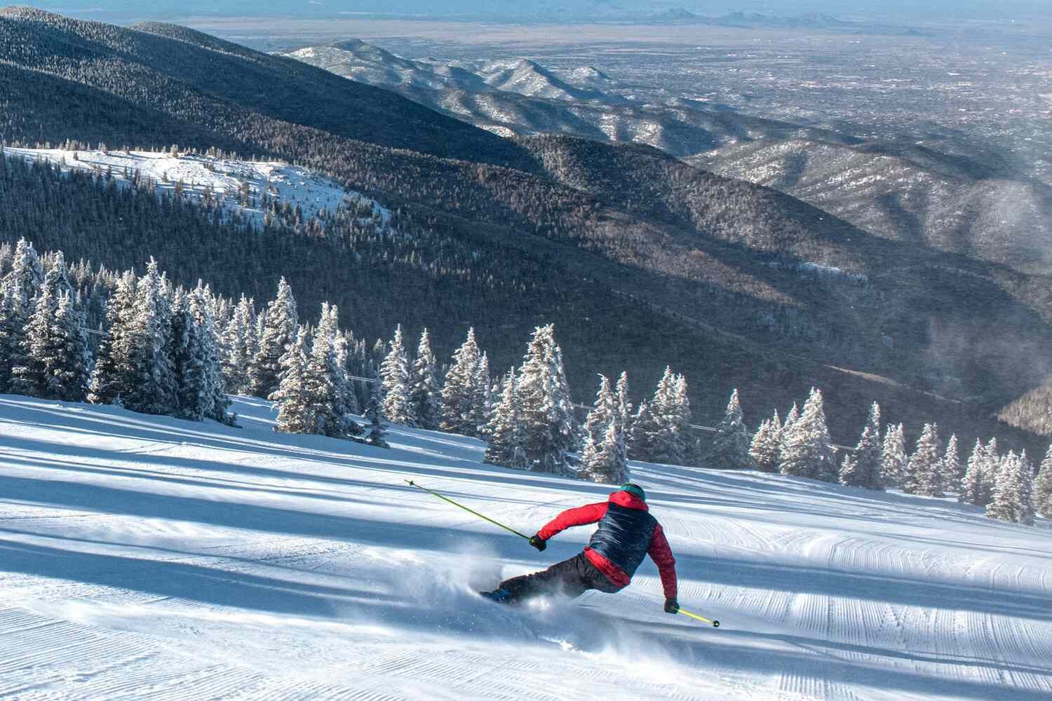 Een skiër in een rode jas skiënd een besneeuwde helling af, met een bebost gebergte op de achtergrond.