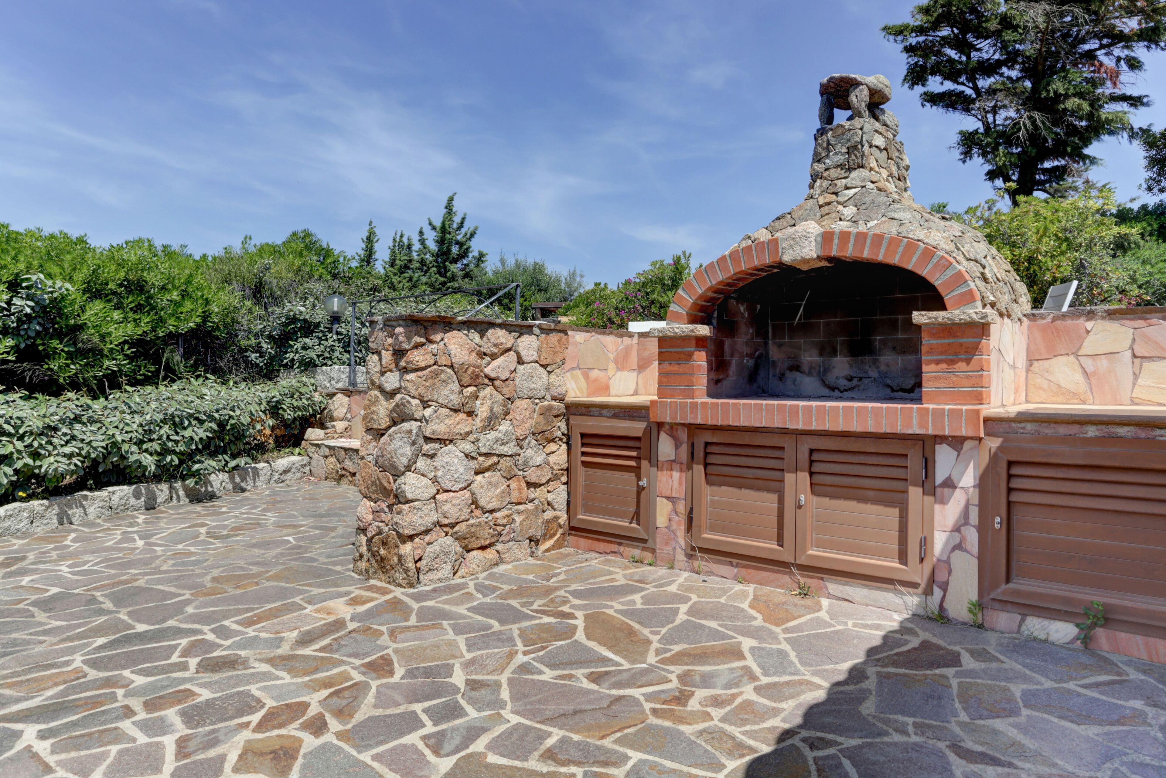 Un patio de piedra que conduce a una cocina al aire libre de ladrillo con chimenea, rodeado de follaje.