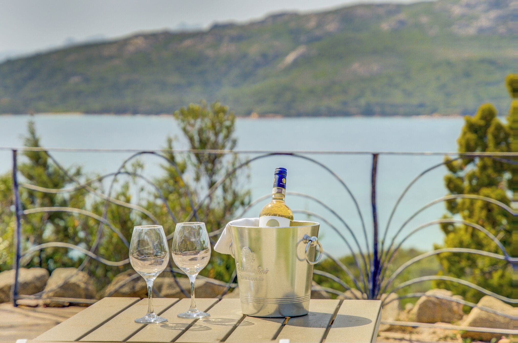 Une table avec une bouteille de champagne et deux verres, sur une terrasse offrant une vue sur un lac et des collines.