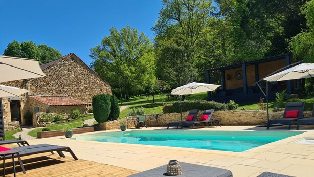 Piscina con sillas de descanso y sombrillas, rodeada de edificios de piedra y vegetación.