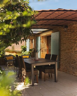 An outdoor dining area with a table and chairs, a stone wall, and a metal roof. - L’Écurie Gîte Two Bedroom (Cassagnes)