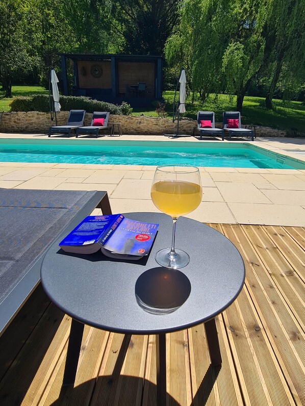 Une table placée au bord de la piscine, avec un livre et un verre, entourée de chaises longues et donnant sur la piscine.