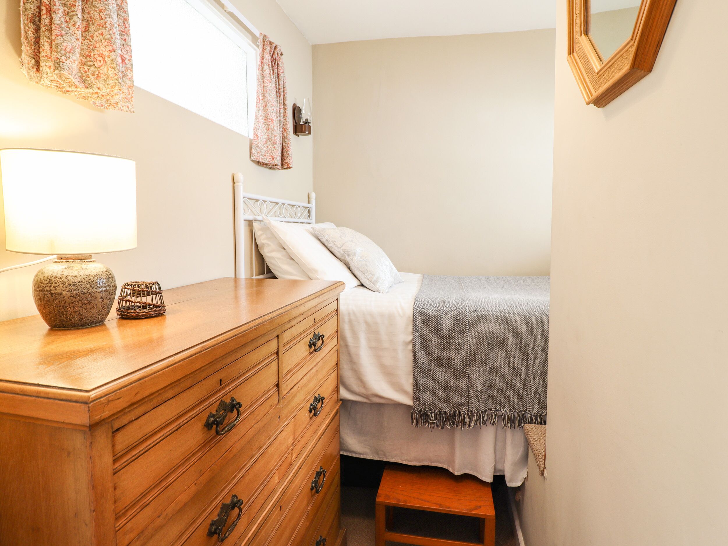 A bedroom with a wooden dresser, a bed with a gray blanket, a nightstand, and a lamp.