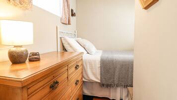 A bedroom with a wooden dresser, a bed with a gray blanket, a nightstand, and a lamp.