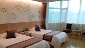 Hotellihuone, jossa on kaksi sänkyä, pieni pöytä, jossa on kuppi ja kasvi, tuoli ja ikkuna, josta on näkymä rakennuksiin.