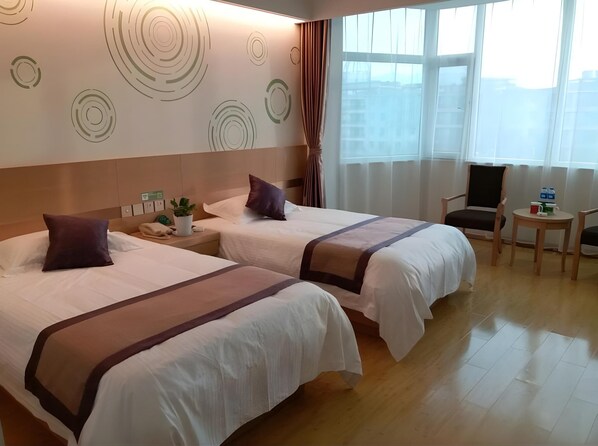 Habitación de hotel con dos camas, una mesita con una taza y una planta, una silla y un ventanal con vistas a edificios.