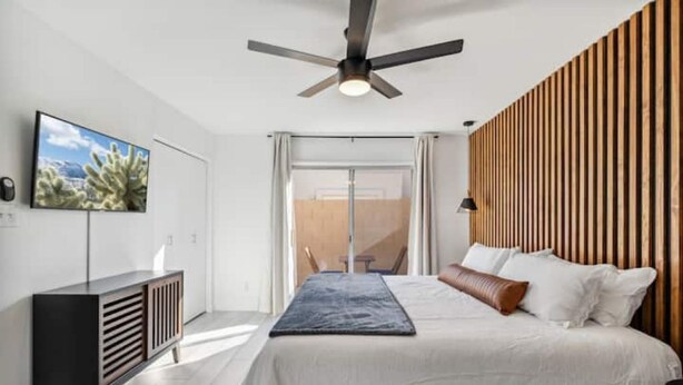 A modern bedroom with a bed, a TV mounted on the wall, a ceiling fan, and a wooden panel wall. - Desert Gem-Walk To Spring Training,ASU,Tempe Lake! (Tempe)