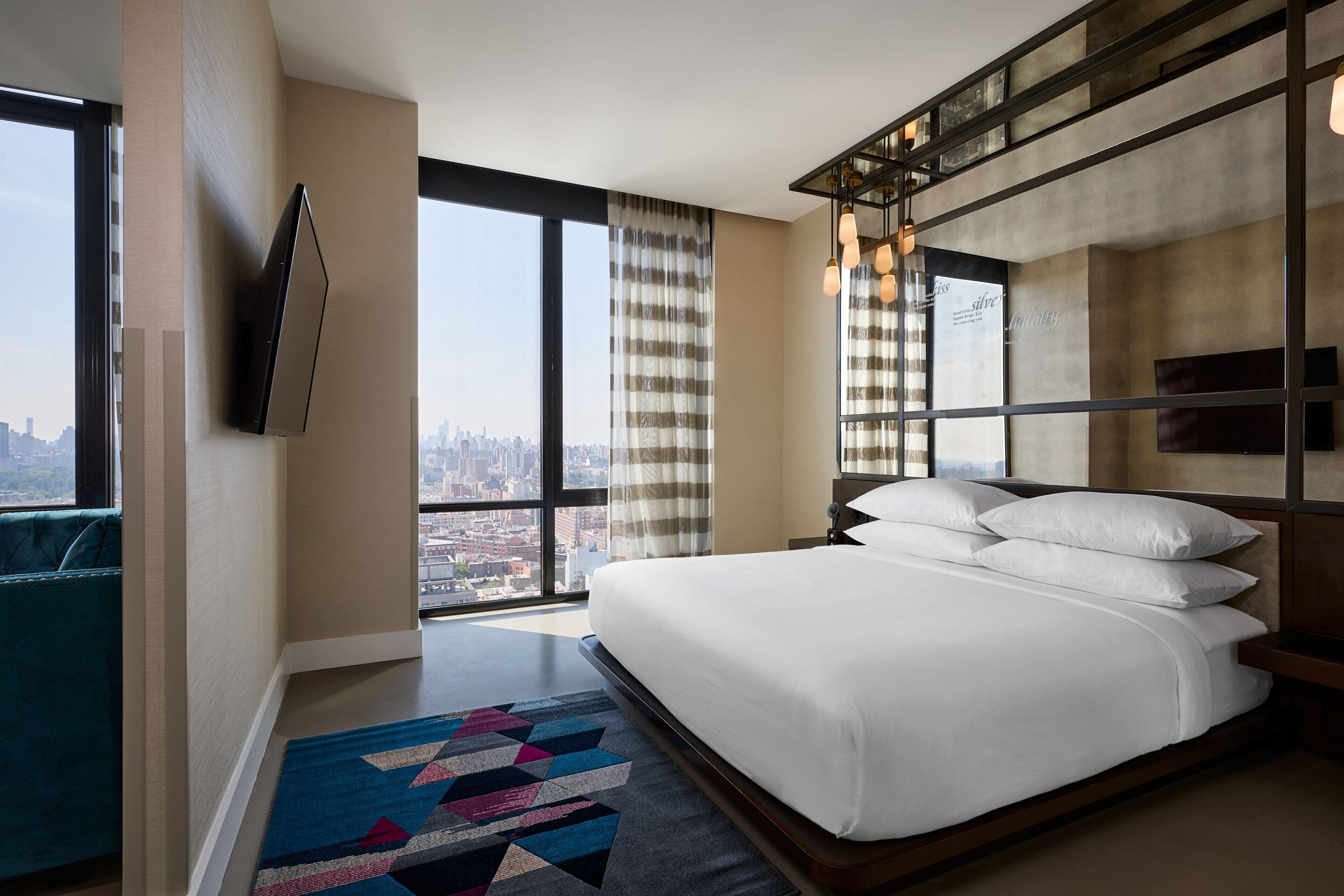 A modern hotel room with a large bed, a city view, a flat-screen TV mounted on the wall, and a unique geometric-patterned rug.