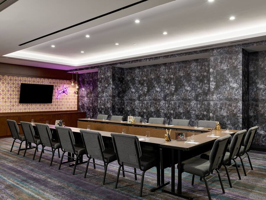 A conference room with a long table, multiple chairs, a flat-screen TV, and a patterned carpet.