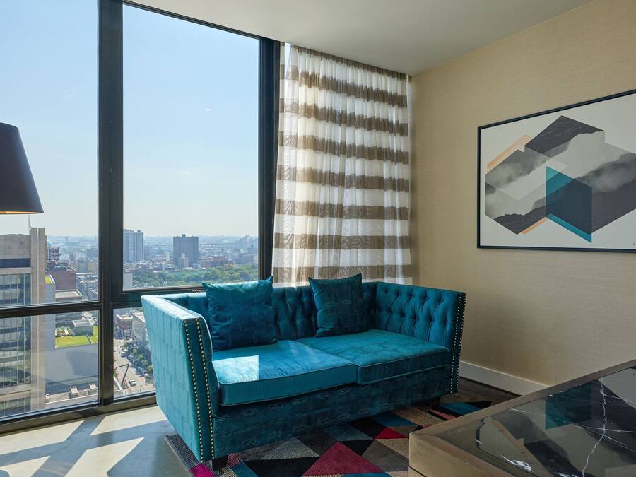 Un salon moderne avec un canapé de couleur turquoise, de grandes fenêtres, une vue sur la ville et une œuvre d’art géométrique accrochée au mur.