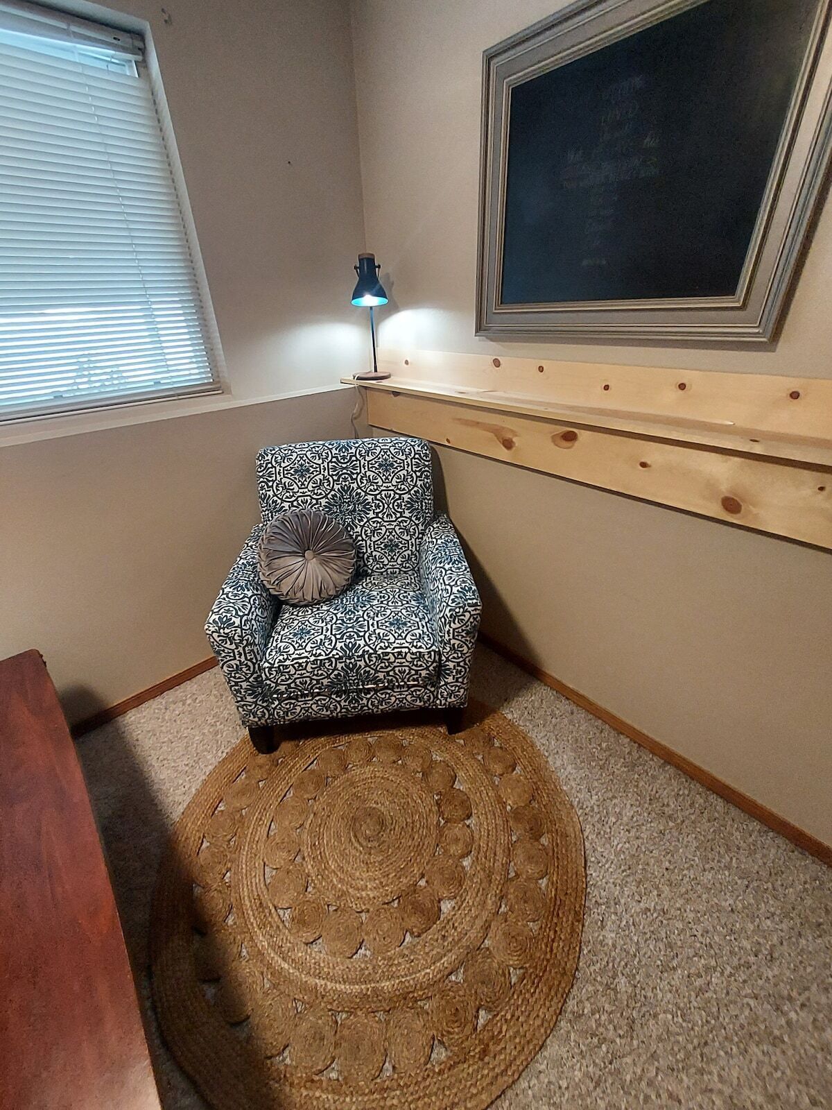 A small, patterned armchair with a cushion, a wooden shelf, a lamp, and a round woven rug in a corner room.