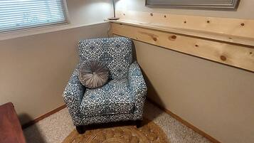 A small, patterned armchair with a cushion, a wooden shelf, a lamp, and a round woven rug in a corner room.